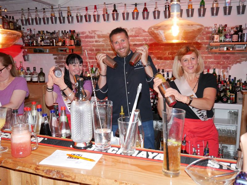 Teilnehmerinnen eines Junggesellinnenabschieds stehen mit Markus hinter der Bar und schütteln Cocktails